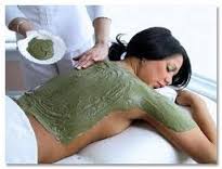 Woman receiving a green body scrub massage on a spa table, with paste being applied to her back